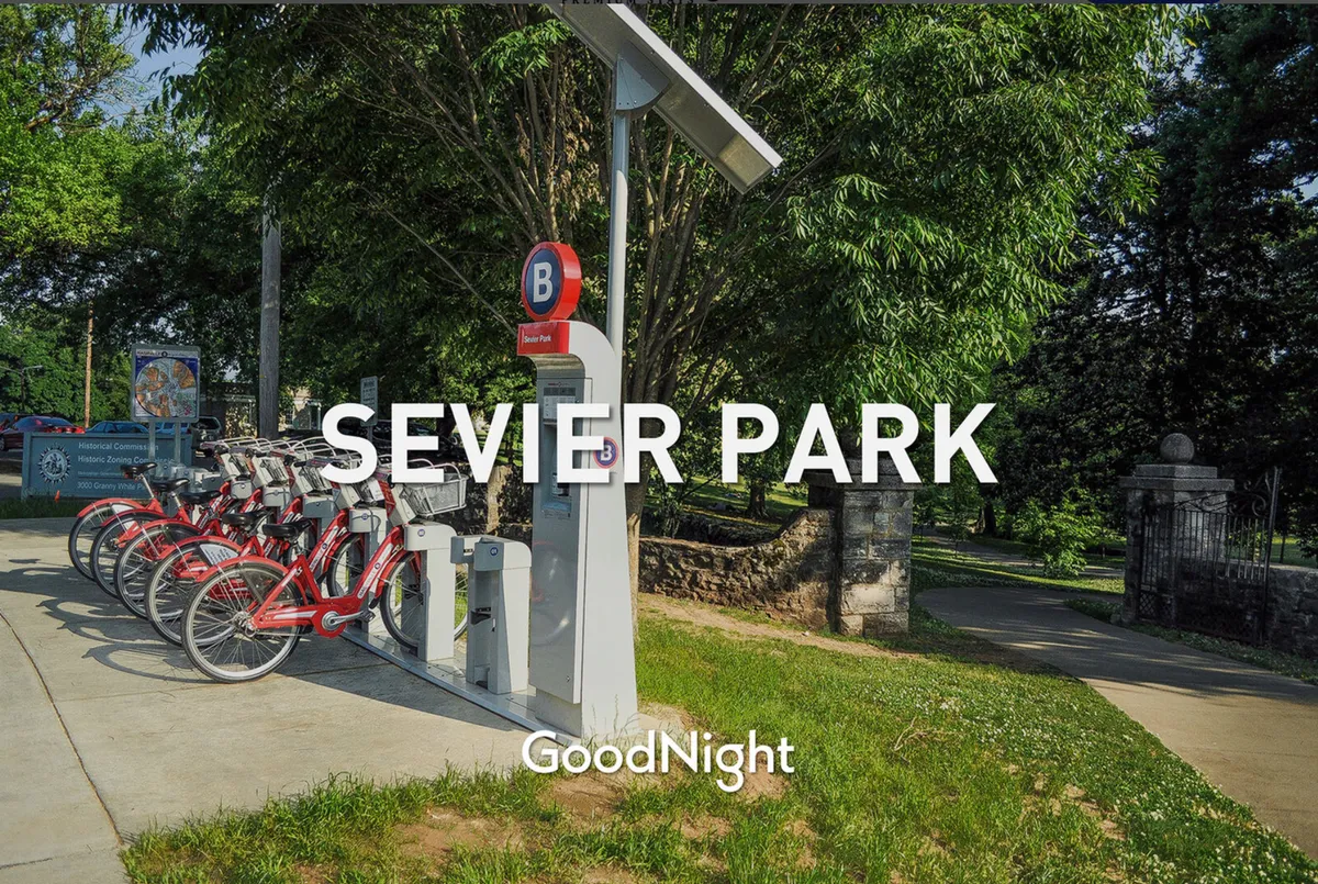 A bike rental station and park entrance set amidst lush greenery, hinting at pathways perfect for walking, biking, or picnics. The inviting park atmosphere connects with local recreational opportunities.