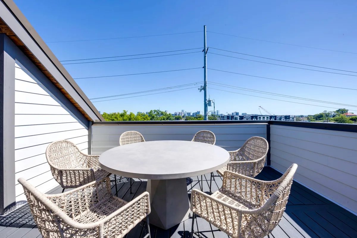 A stylish outdoor dining space with a round concrete table and woven chairs set against the backdrop of the Nashville city skyline. The scene captures the mood of social gatherings and relaxation, with the city’s lights beginning to come alive at dusk.