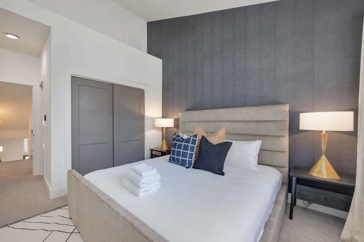 A tranquil bedroom with a plush headboard, decorative pillows, nightstands with lamps, and closet doors. Soft natural light from the windows creates a restful, welcoming environment.