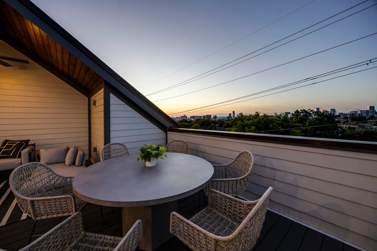 A relaxed outdoor dining setup with a round concrete table, woven chairs, and a sunset sky. The distant city skyline completes the scene, suggesting a peaceful yet social environment for al fresco meals or small celebrations.