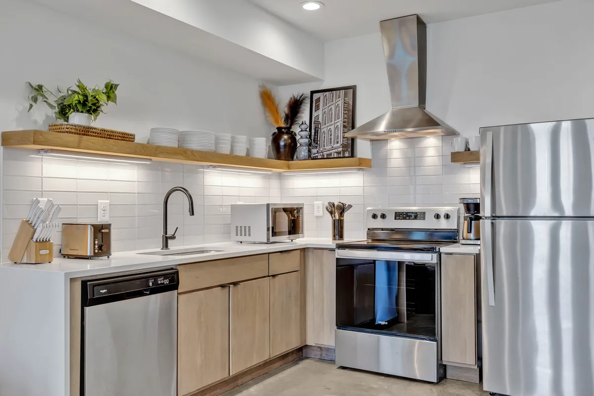 A contemporary kitchen with sleek stainless steel appliances, open wooden shelves, a decorative vase, and a white tile backsplash. The scene is warm and inviting, ideal for hosting, baking, or intimate breakfasts.