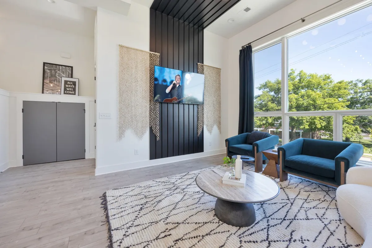 A stylish living space featuring a large window letting in abundant sunlight, a modern TV feature wall, plush seating, and a decorative area rug. The arrangement of seats encourages conversation and relaxation, with a Smart TV positioned for easy viewing.